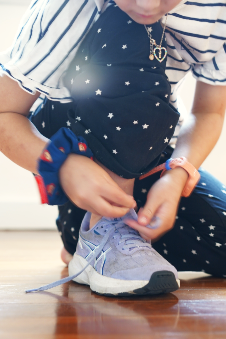 A child kneeling on a wooden floor, focused on tying their purple sneakers. They are wearing a striped shirt and star-patterned leggings, capturing a moment of developing independence.